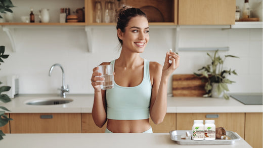 Smiling woman in a modern kitchen holding a glass of water and a supplement capsule, with Kardish brand supplement bottles on a tray in the foreground, promoting health and wellness.